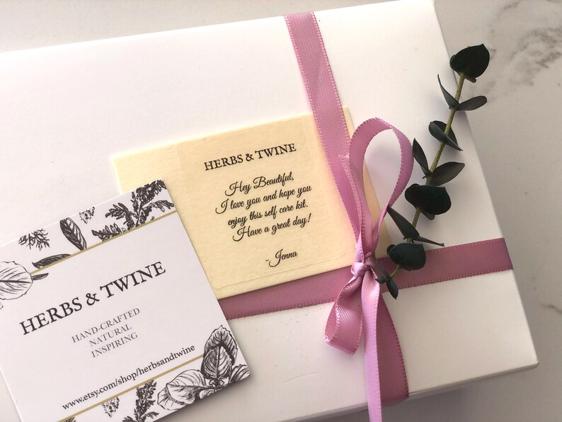 May include: A white gift box with a pink ribbon and a small sprig of eucalyptus. The box is tied with a pink ribbon and has a small card attached to it that says "HERBS & TWINE" and "Hey Beautiful, I love you and hope you enjoy this self care kit. Have a great day! - Jenna".
