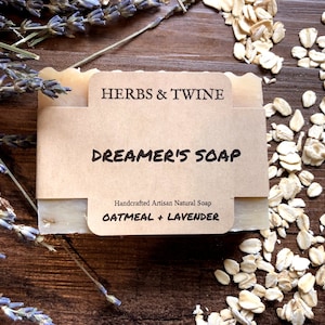 May include: A bar of Dreamer's Soap with a brown paper label that reads "HERBS & TWINE" and "DREAMER'S SOAP". The soap is surrounded by dried lavender and oats on a wooden surface. The label also says "Oatmeal + Lavender".