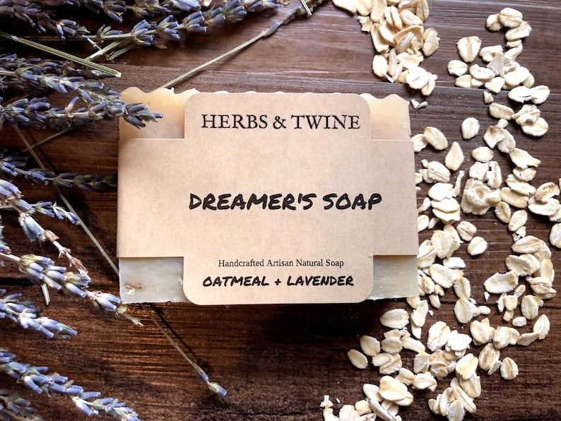 May include: A bar of Dreamer's Soap with a brown paper label that reads "HERBS & TWINE" and "DREAMER'S SOAP". The soap is surrounded by dried lavender and oats on a wooden surface. The label also says "Oatmeal + Lavender".