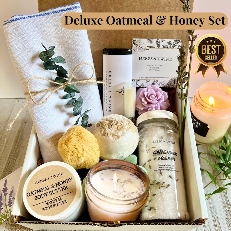 May include: A gift set labeled "Deluxe Oatmeal & Honey Set" includes a white towel, bath bombs, a candle, body butter, and a jar of bath salts. The set contains a sponge, a bar of soap, and a decorative flower. The box is decorated with sprigs of greenery.