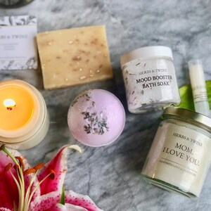 May include: A gift set for mothers with a lavender bath bomb, a candle, a bar of soap, a jar of bath salts, and a small jar of body butter. The bath salts jar has the text "Herbs & Twine Mood Booster Bath Soak" and the body butter jar has the text "Herbs & Twine Mom, I Love You. Hand with Love & Gratitude."