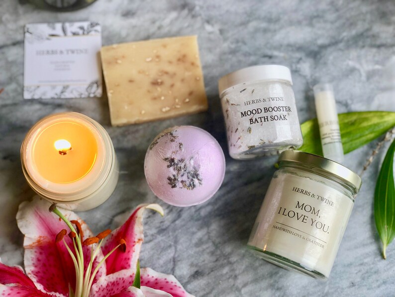 May include: A gift set for mothers with a lavender bath bomb, a candle, a bar of soap, a jar of bath salts, and a small jar of body butter. The bath salts jar has the text "Herbs & Twine Mood Booster Bath Soak" and the body butter jar has the text "Herbs & Twine Mom, I Love You. Hand with Love & Gratitude."