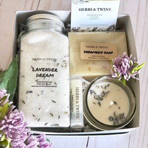 May include: A gift box filled with lavender-themed bath and body products. The box contains a jar of lavender bath salts, a bar of oatmeal and lavender soap, a small jar of lavender candle, and a lavender lip balm. The box is decorated with a small card that says "Herbs & Twine" and a sprig of lavender.