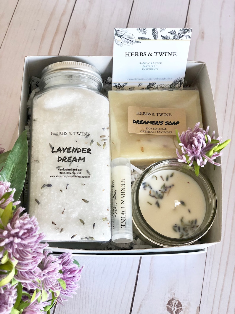 May include: A gift box filled with lavender-themed bath and body products. The box contains a jar of lavender bath salts, a bar of oatmeal and lavender soap, a small jar of lavender candle, and a lavender lip balm. The box is decorated with a small card that says "Herbs & Twine" and a sprig of lavender.