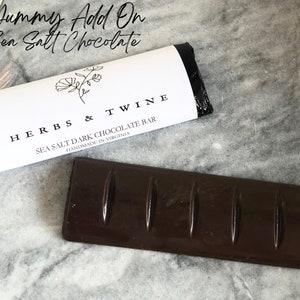 May include: A dark chocolate bar with sea salt, wrapped in black and white packaging with the text "HERBS & TWINE SEA SALT DARK CHOCOLATE BAR HANDMADE IN VIRGINIA".