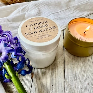 May include: A jar of Oatmeal & Honey Body Butter with a label that reads "Herbs & Twine". A lit candle in a gold tin and purple flowers are also in the image. The body butter is labeled "Natural Body Butter".