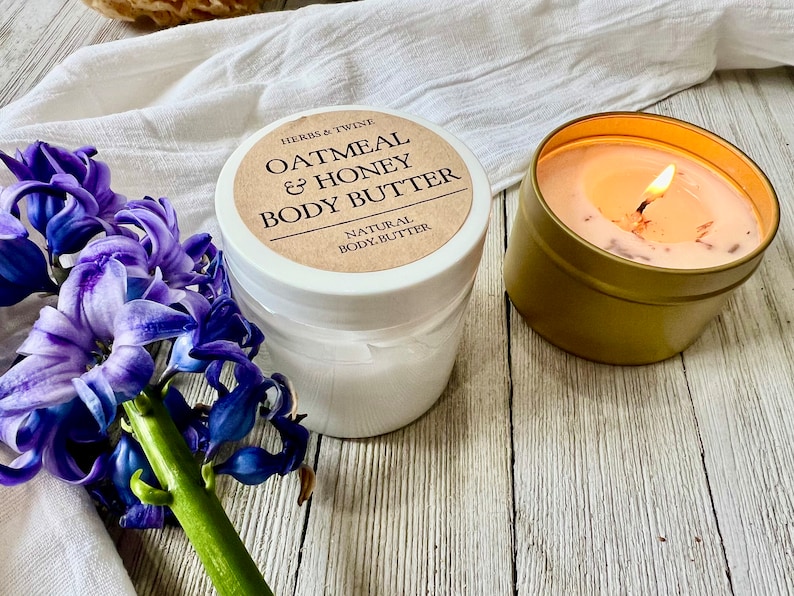 May include: A jar of Oatmeal & Honey Body Butter with a label that reads "Herbs & Twine". A lit candle in a gold tin and purple flowers are also in the image. The body butter is labeled "Natural Body Butter".
