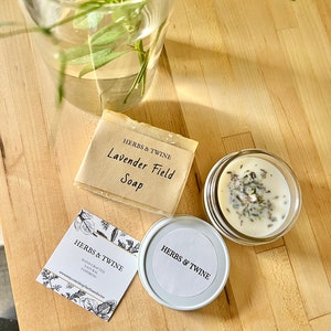 May include: A set of natural bath and body products from Herbs & Twine. The set includes a bar of lavender field soap, a small jar of candle with dried lavender, and a business card with the company's logo and website.