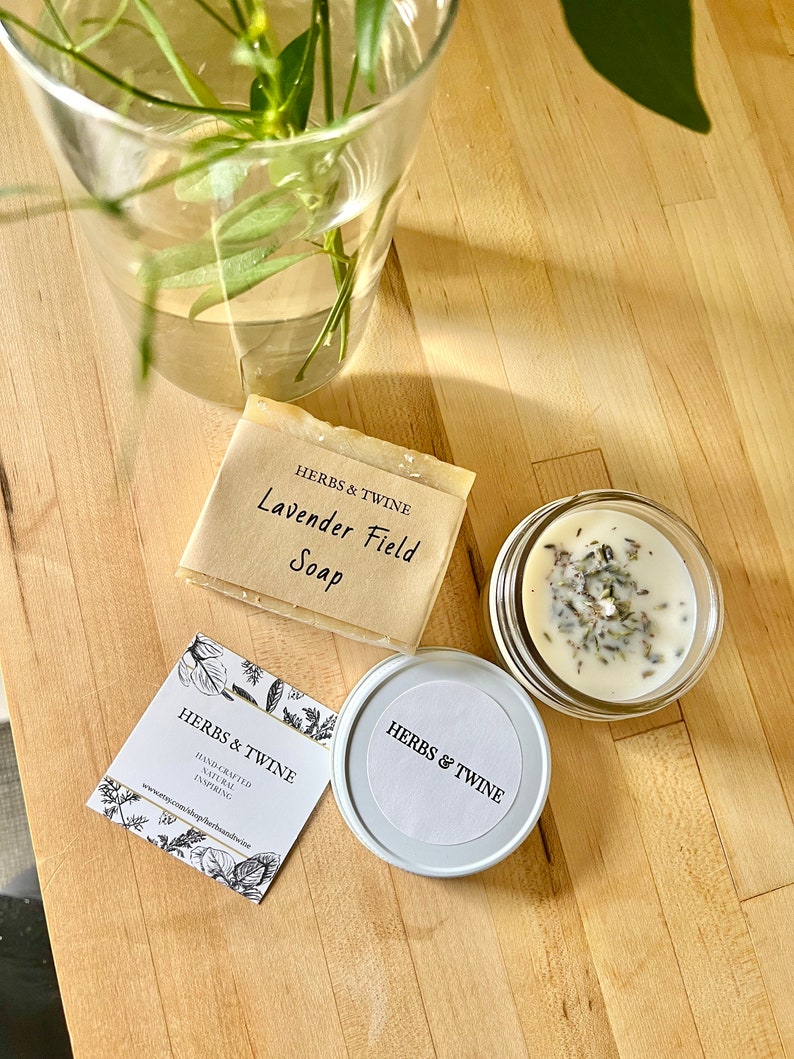 May include: A set of natural bath and body products from Herbs & Twine. The set includes a bar of lavender field soap, a small jar of candle with dried lavender, and a business card with the company's logo and website.