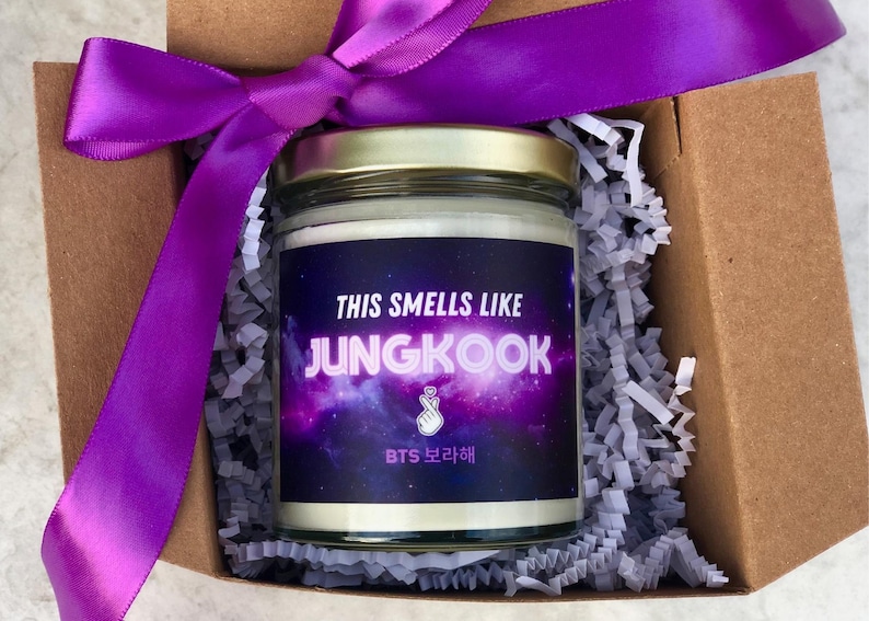 Puede incluir: Un tarro de vidrio para velas con una etiqueta morada con tem&aacute;tica de galaxia que dice "This smells like Jungkook BTS 보라해". El tarro est&aacute; en una caja de cart&oacute;n marr&oacute;n con cinta morada y papel triturado.