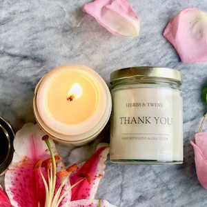 May include: A white candle in a glass jar with a gold lid. The candle is lit and the flame is visible. The jar has a label that says "HERBS & TWINE" and "THANK YOU" with the text "HANDWITH LOVE & GRATITUDE" below. The candle is surrounded by pink rose petals and a pink lily.
