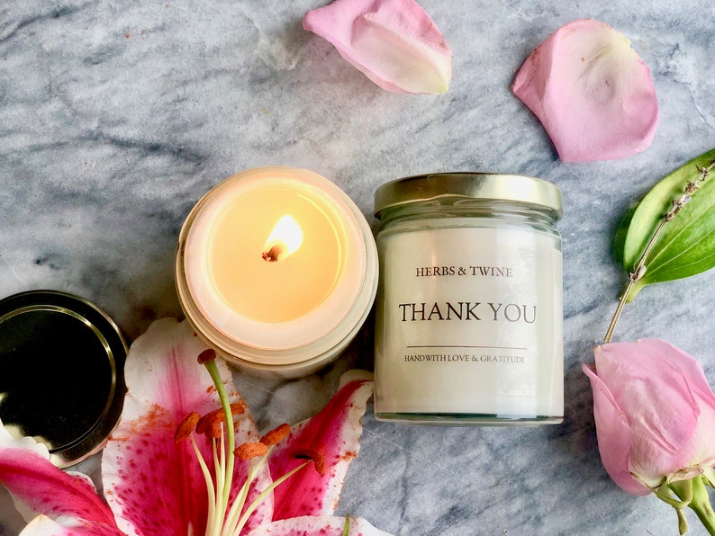 May include: A white candle in a glass jar with a gold lid. The candle is lit and the flame is visible. The jar has a label that says "HERBS & TWINE" and "THANK YOU" with the text "HANDWITH LOVE & GRATITUDE" below. The candle is surrounded by pink rose petals and a pink lily.