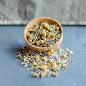 May include: A wooden bowl filled with dried chamomile and lavender flowers. The bowl is tipped over, spilling the flowers onto a gray surface.