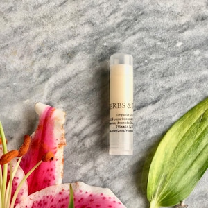 Può includere: Un tubo trasparente di balsamo per labbra con un'etichetta bianca che recita "HERBS &" e elenca gli ingredienti, tra cui "Organic Shea", "100% pure Coconut", "Avocado Oil" e "Vitamin E".