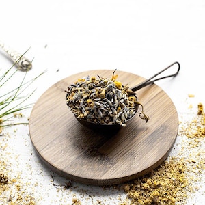 May include: A metal tea infuser filled with dried herbs and flowers, resting on a wooden cutting board. The infuser is surrounded by loose herbs and a sprig of green foliage, suggesting a natural, herbal tea blend.
