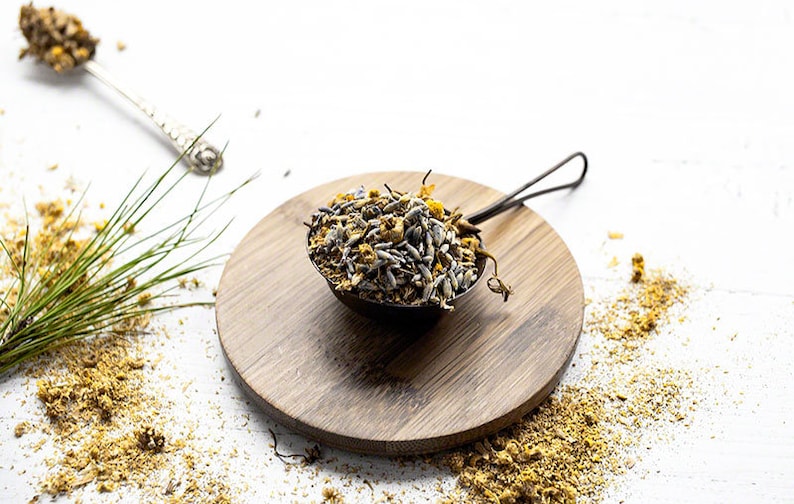 May include: A metal tea infuser filled with dried herbs and flowers, resting on a wooden cutting board. The infuser is surrounded by loose herbs and a sprig of green foliage, suggesting a natural, herbal tea blend.