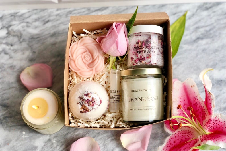 May include: A gift box filled with bath and body products, including a bath bomb, soap, bath salts, and a candle. The box is decorated with shredded paper and a pink rose. The products are labeled with the text "Mood Booster Bath Soak" and "Thank You".
