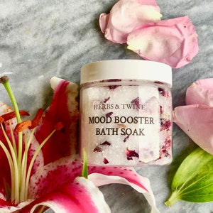 May include: A clear jar filled with white bath salts and pink rose petals. The jar has a white lid and a label that reads "Herbs & Twine Mood Booster Bath Soak".