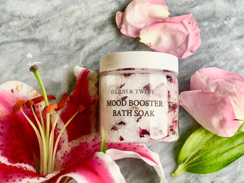 May include: A clear jar filled with white bath salts and pink rose petals. The jar has a white lid and a label that reads "Herbs & Twine Mood Booster Bath Soak".