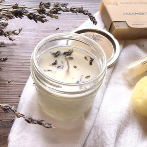 May include: A clear glass jar candle with lavender buds, a white wick, and a metal lid. The candle is next to a bar of soap with the text "Dreamer's Soap" and a lip balm tube. Dried lavender sprigs are also present.