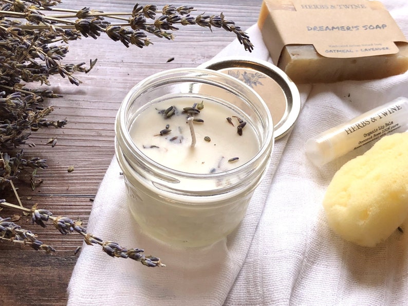 May include: A clear glass jar candle with lavender buds, a white wick, and a metal lid. The candle is next to a bar of soap with the text "Dreamer's Soap" and a lip balm tube. Dried lavender sprigs are also present.