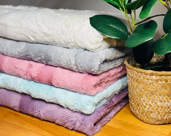 May include: A stack of plush, faux fur blankets in various pastel colors: white, gray, pink, light blue, and lavender. The blankets have a soft, textured appearance and are neatly folded. A decorative plant in a woven basket is in the background.