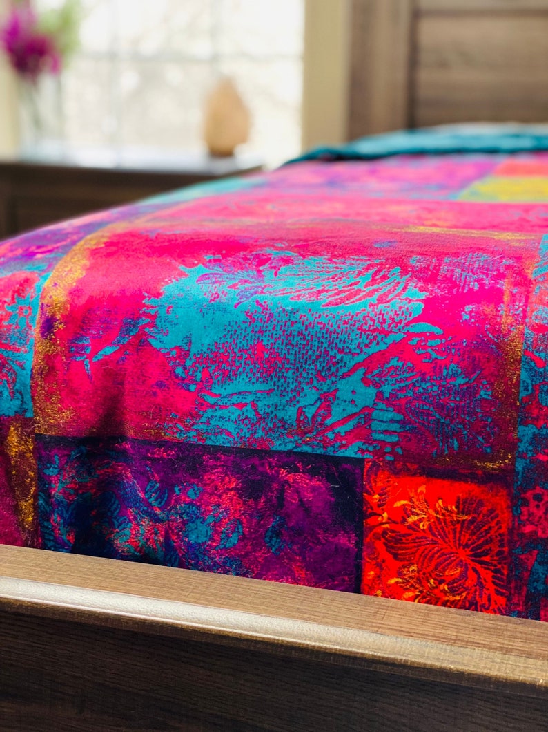 May include: A close-up of a pink, teal, purple, and red patterned bedspread. The bedspread is made of a soft, plush fabric and has a geometric design.