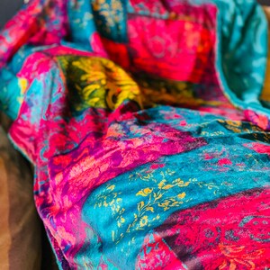 May include: A colorful throw blanket with a patchwork design featuring teal, pink, purple, and gold hues. The blanket has a soft, fuzzy texture.