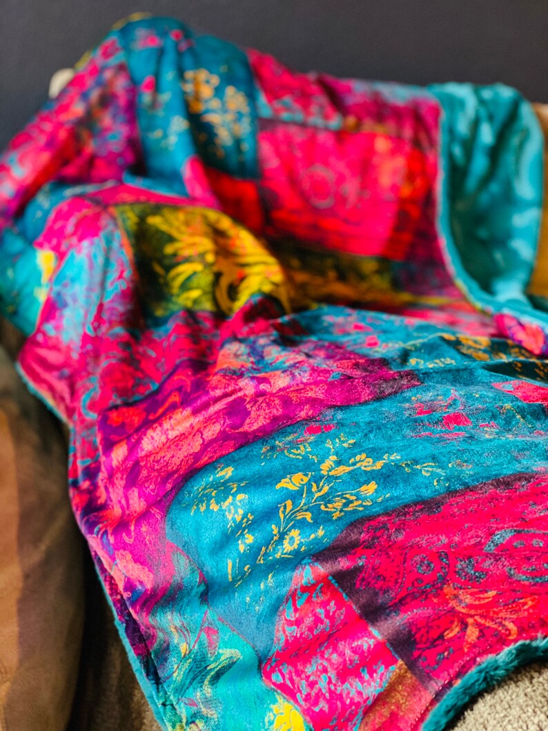 May include: A colorful throw blanket with a patchwork design featuring teal, pink, purple, and gold hues. The blanket has a soft, fuzzy texture.