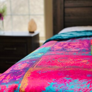 May include: A close-up of a colorful patterned bedspread with shades of pink, blue, yellow, and purple. The bedspread is made of a soft, textured fabric and features a floral design.