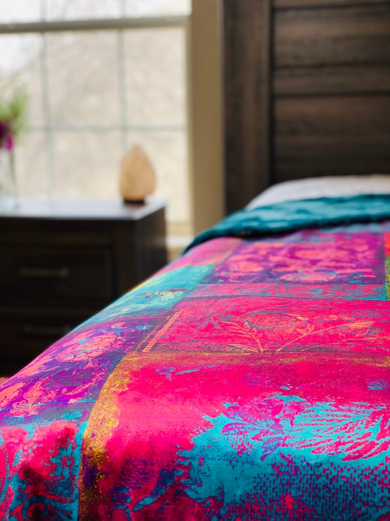 May include: A close-up of a colorful patterned bedspread with shades of pink, blue, yellow, and purple. The bedspread is made of a soft, textured fabric and features a floral design.