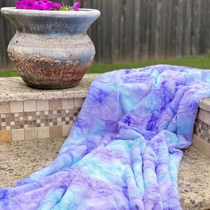 May include: A plush, faux fur blanket in shades of blue and purple, draped over a stone surface. The blanket has a soft, textured appearance. A decorative, weathered ceramic pot with purple flowers sits nearby.