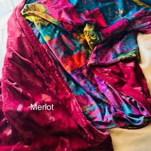 May include: A plush throw blanket with a patchwork design in shades of red, blue, green, and yellow. The blanket is folded and draped over a white surface. The text "Merlot" is visible on the blanket.