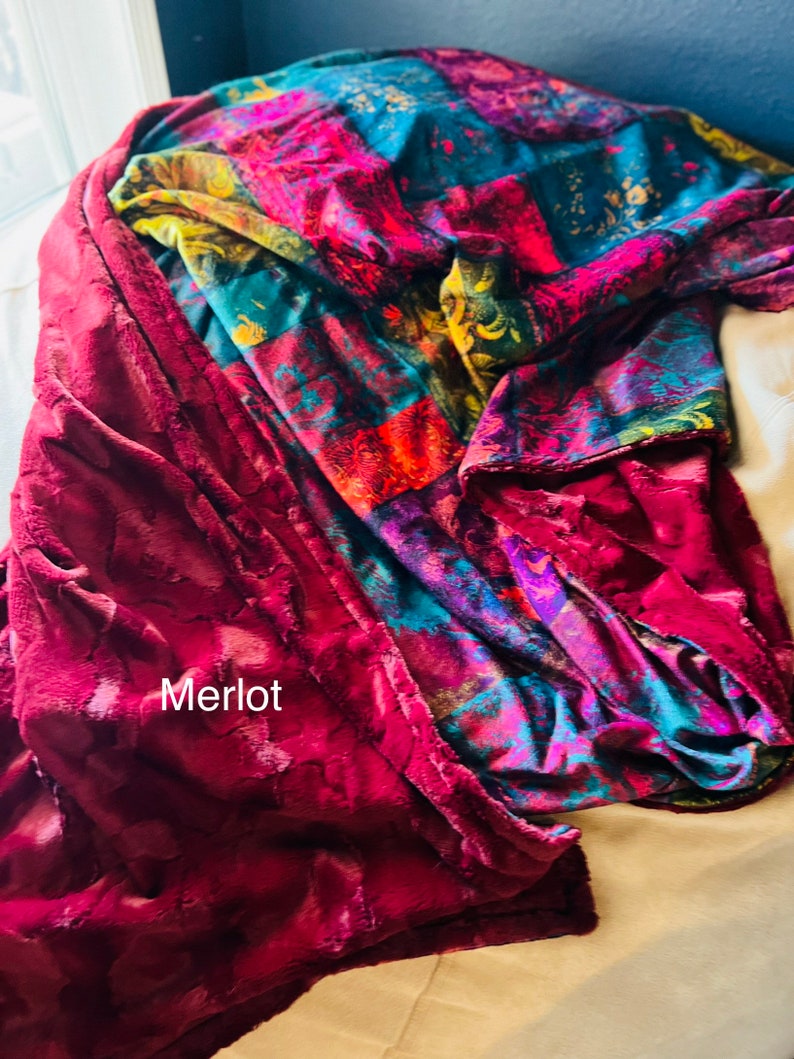 May include: A plush throw blanket with a patchwork design in shades of red, blue, green, and yellow. The blanket is folded and draped over a white surface. The text "Merlot" is visible on the blanket.