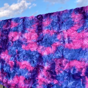 May include: A plush, faux fur blanket in shades of blue, purple, and pink. The blanket has a textured, rippled appearance, with the colors blending in a tie-dye or watercolor effect. The blanket is draped over a railing against a blue sky.