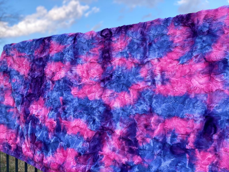 May include: A plush, faux fur blanket in shades of blue, purple, and pink. The blanket has a textured, rippled appearance, with the colors blending in a tie-dye or watercolor effect. The blanket is draped over a railing against a blue sky.