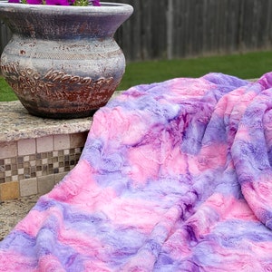 May include: A plush, tie-dye blanket in shades of pink and purple. The soft, textured blanket is draped over a surface, with a decorative flower pot in the background. The blanket's colors create a dreamy, watercolor effect.