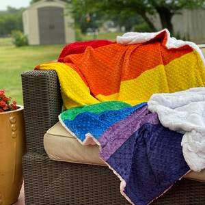 May include: A rainbow-colored plush blanket with a white faux fur backing. The blanket is folded and draped over a wicker couch.