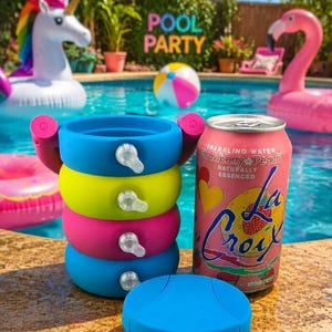 May include: A stack of four colourful silicone beverage holders in blue, yellow, and pink, each with a small clear tab, positioned next to a pink LaCroix Strawberry Peach sparkling water can and a separate blue circular silicone lid.