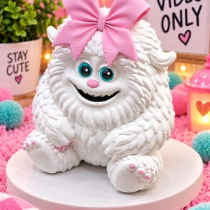 May include: A white, furry creature figurine with a large pink bow on its head, teal eyes, and a small pink nose. The creature has a wide, smiling mouth, textured fur detailing, and small pink accents on its paws, presented as a decorative item.
