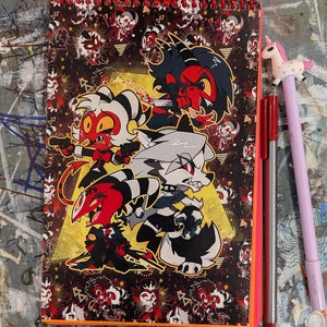 May include: Spiral-bound notebook featuring a colorful cartoon illustration of several characters. The cover has a black background with a repeating pattern of the characters. Two red pens and a unicorn pen are next to the notebook.
