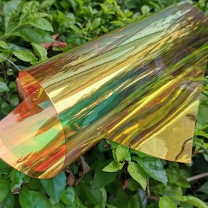A rolled sheet of iridescent film, displaying a spectrum of colors including gold, green, and orange. The film is partially unrolled, revealing its reflective and translucent qualities. The background features green foliage.