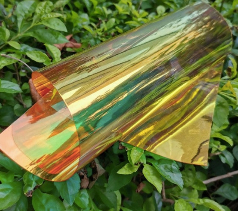A rolled sheet of iridescent film, displaying a spectrum of colors including gold, green, and orange. The film is partially unrolled, revealing its reflective and translucent qualities. The background features green foliage.