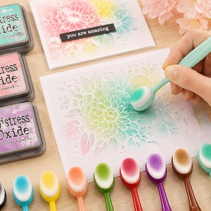 May include: A craft scene featuring Distress Oxide ink pads in teal, pink, and purple, along with a variety of colorful blending brushes. A hand uses a brush to apply ink to a floral stencil. The text "you are amazing" is on a black label.