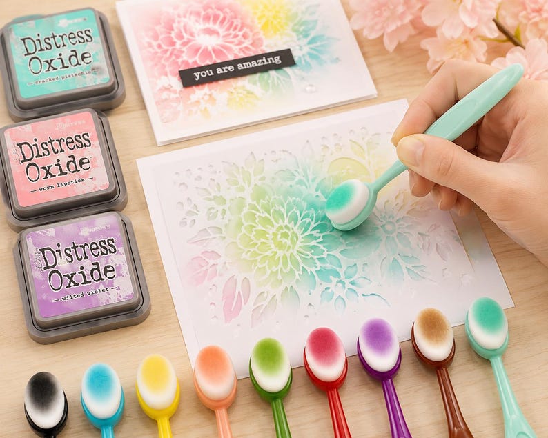 May include: A craft scene featuring Distress Oxide ink pads in teal, pink, and purple, along with a variety of colorful blending brushes. A hand uses a brush to apply ink to a floral stencil. The text "you are amazing" is on a black label.