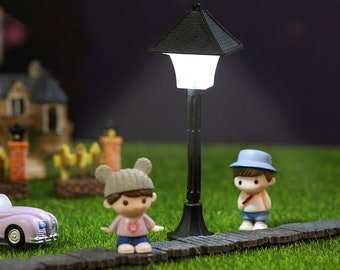 May include: A miniature black street lamp with a white light illuminates a miniature scene with two figures and a car. The lamp is on a path made of small gray stones.