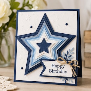 Puede incluir: Tarjeta de cumpleaños hecha a mano con un diseño de estrella en capas en tonos azules y blancos. La tarjeta incluye el texto "Happy Birthday" en una pancarta, adornada con hojas y un lazo de cuerda. Fondo blanco texturizado.