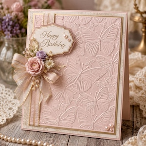 Puede incluir: Una tarjeta de cumpleaños rosa pálido con un patrón de mariposas en relieve, una etiqueta con borde dorado con el texto Happy Birthday, un lazo de cinta transparente y pequeñas flores de tela, decorada con perlas blancas.