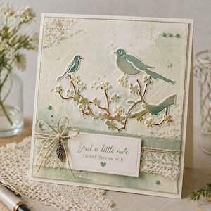 May include: A handmade greeting card featuring two teal birds perched on a branch with white flowers. The card has a textured, off-white background with a decorative ribbon and a tag that reads "Just a little note to say thank you."