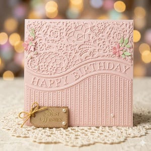 Może przedstawiać: R&oacute;żowa kartka urodzinowa z wytłoczonym napisem "Happy Birthday" na froncie. Karta ma dekoracyjny wz&oacute;r przypominający koronkę i jest ozdobiona małymi r&oacute;żowymi kwiatami i perłami. Dołączona jest mała etykieta z napisem "Best Wishes".
